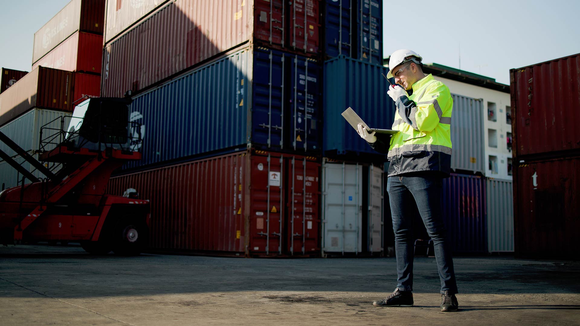 Man with tablet communicating with someone over a walkie talkie with cargo containers in the background