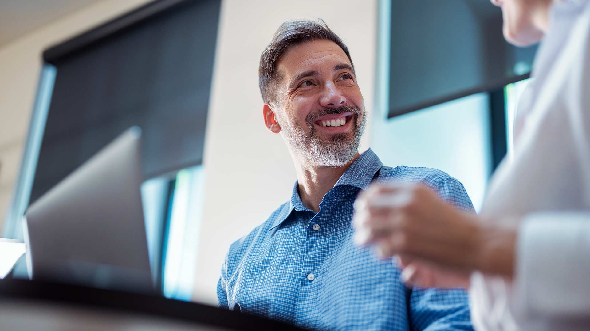 Man smiling at desk in front of laptop talking with someone nearby